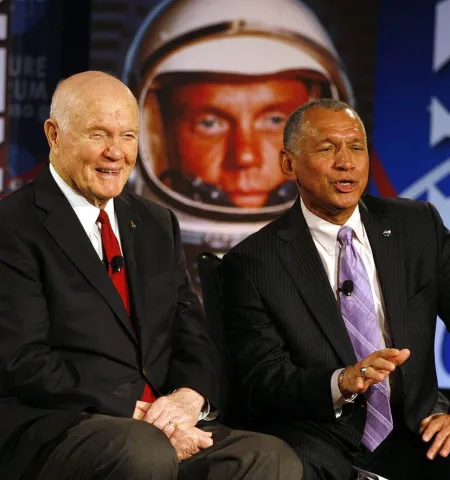 Sen. John Glenn sitting beside Maj. Gen. Charles F. Bolden Jr. with a photo of John Glenn's face in astronaut helmet in background