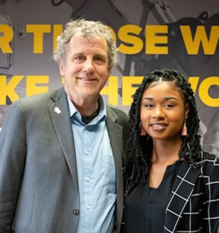 a man and a woman stand inside in front of a wall with the words “for those who make the world move” on it.