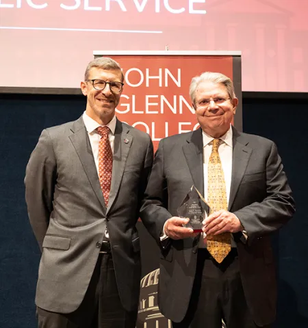 Glenn College Dean Trevor Brown, left, and U.S. Comptroller General Gene Dodaro stand on a stage. Dodaro holds the crystal glass John Glenn Excellence in Public Service award and, in the background, is a partially obscured sign that reads John Glenn College of Public Affairs.