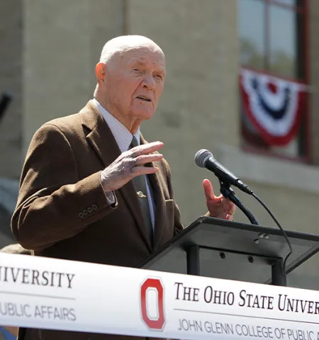 Senator Glenn moving his hands as he speaks to a crowd behind a microphone stand.