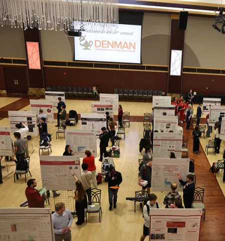 In a large room, students stand by research posters and discuss them with judges and visitors.